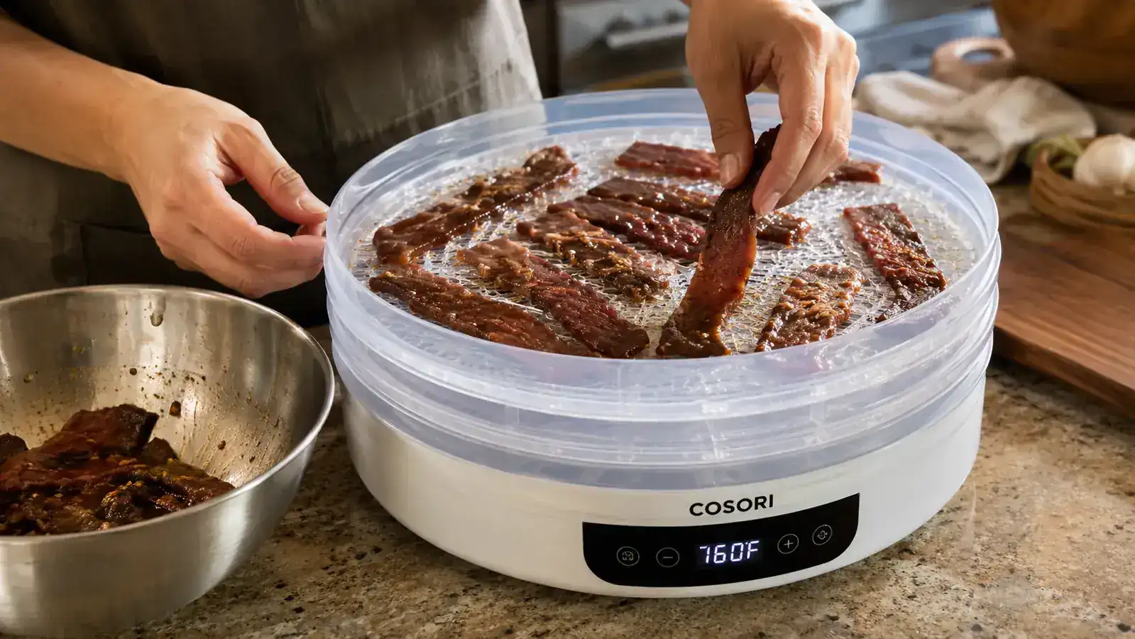 Hand loading thin strips of marinated beef onto a dehydrator tray next to the Cosori unit