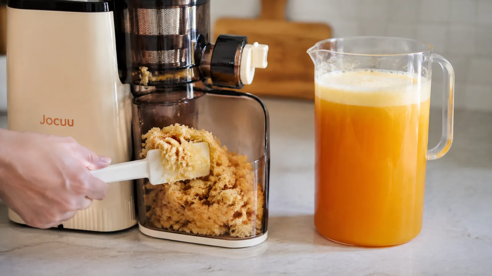 Dry, crumbly pulp being scraped from the Jocuu juicer's pulp bin next to a nearly empty juice pitcher