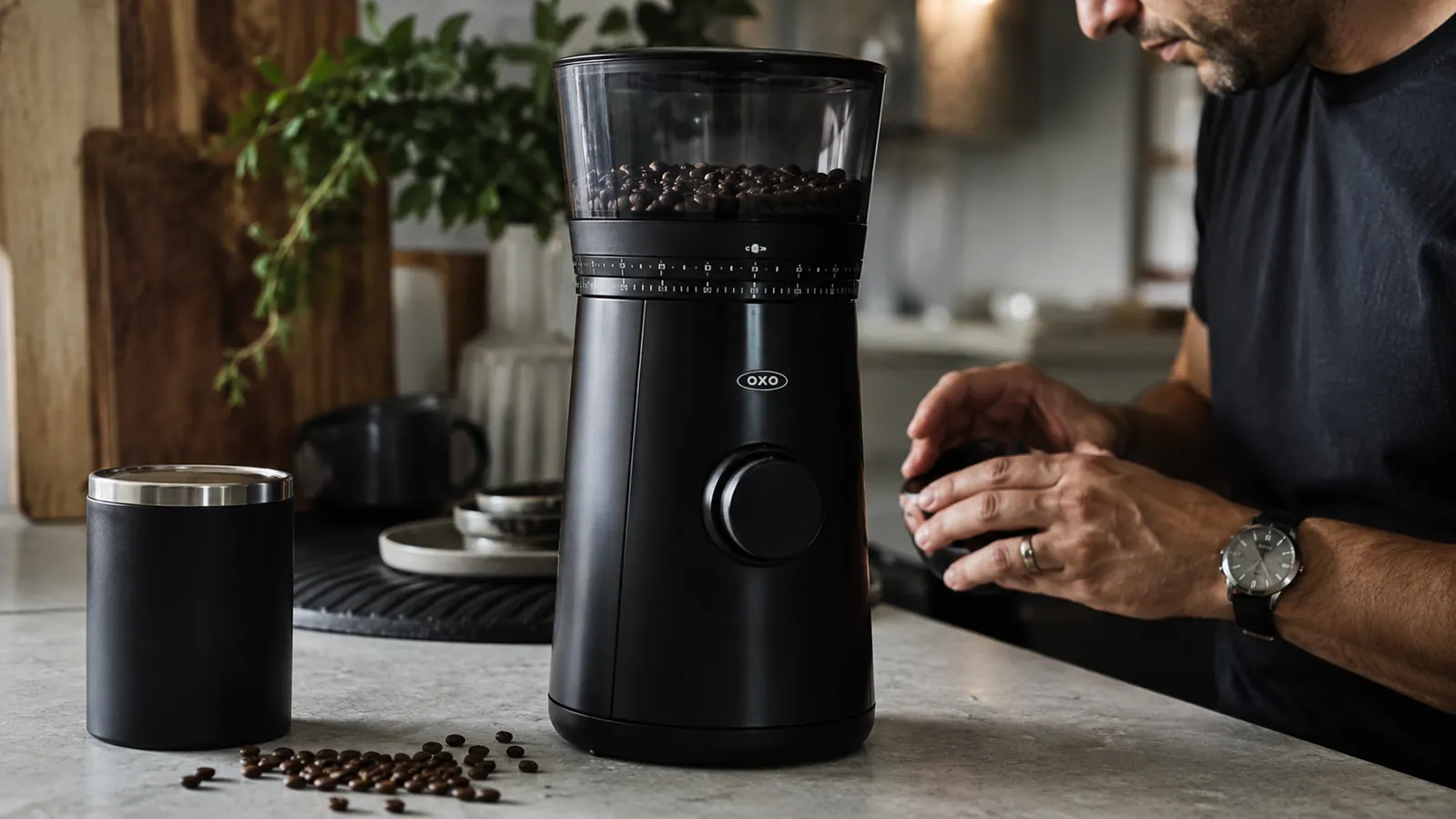 Matte black OXO Brew Conical Burr Grinder on a kitchen counter with loose coffee beans scattered around the base