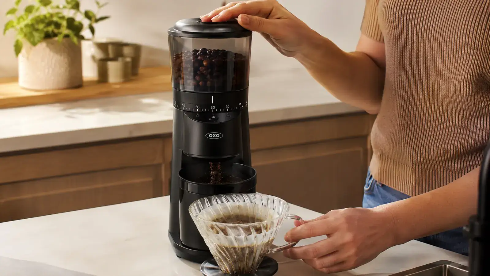A hand pressing the one-touch button on the OXO grinder while ground coffee falls into a pour-over filter