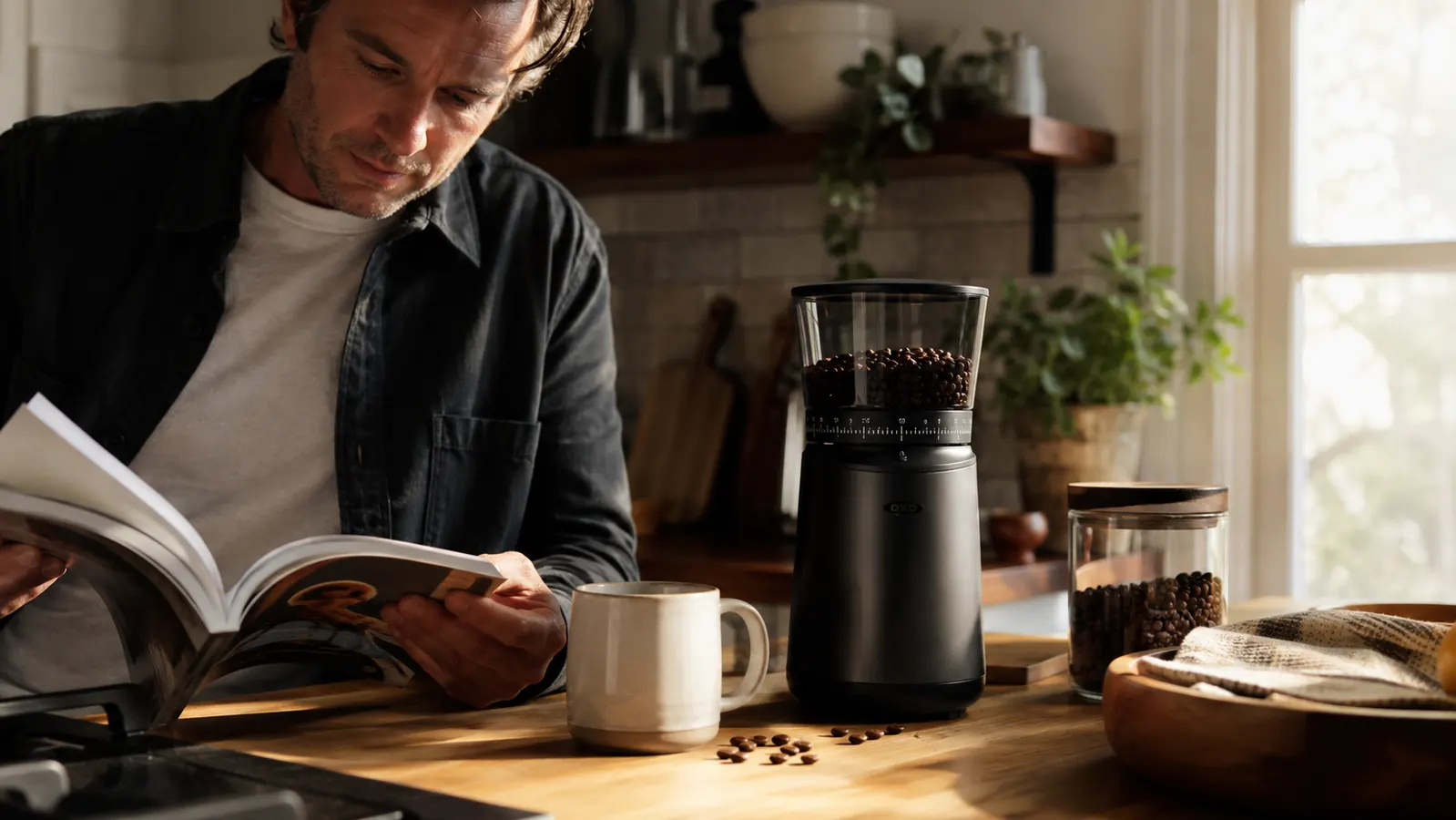 Matte black OXO Brew Conical Burr Grinder sitting on a kitchen counter in soft morning light, beans scattered nearby
