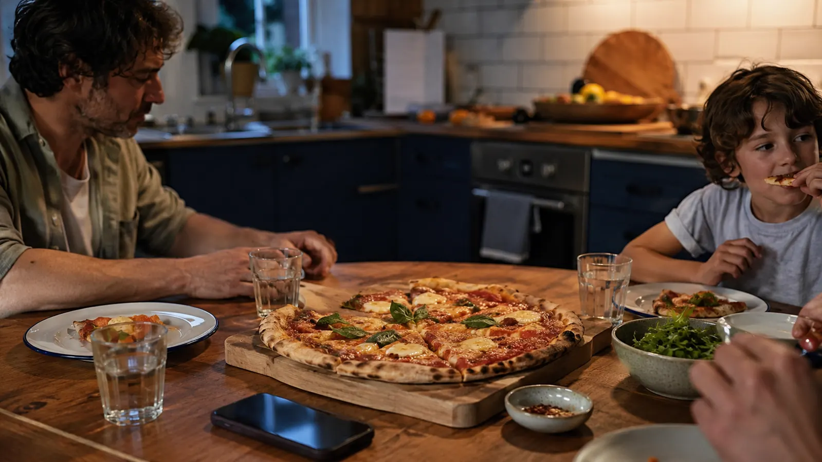 A family kitchen table set for pizza night with plates, a cut pizza, and a phone with a delivery app closed beside it