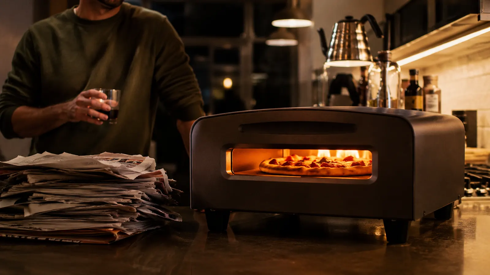 Countertop pizza oven glowing on a kitchen counter at dusk with a stack of old delivery menus set beside it